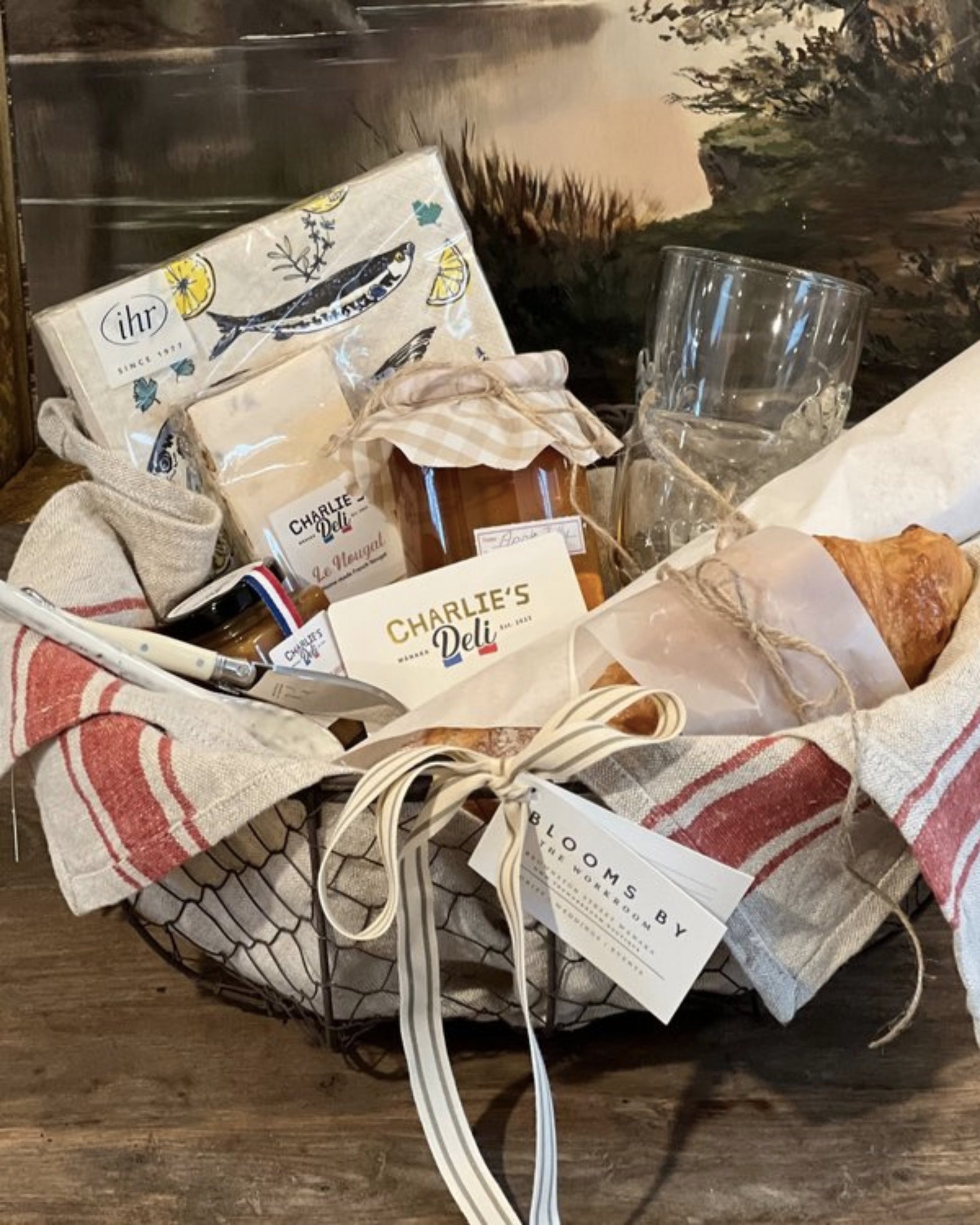 Introducing The Workroom's French Lovers Picnic Gift Basket: a charming collection that includes an artisanal homemade fruit spread, a croissant wrapped in paper, tea towels, a candle, and a glass tumbler. Elegantly tied with ribbon and featuring business cards from Charlie's French Deli.