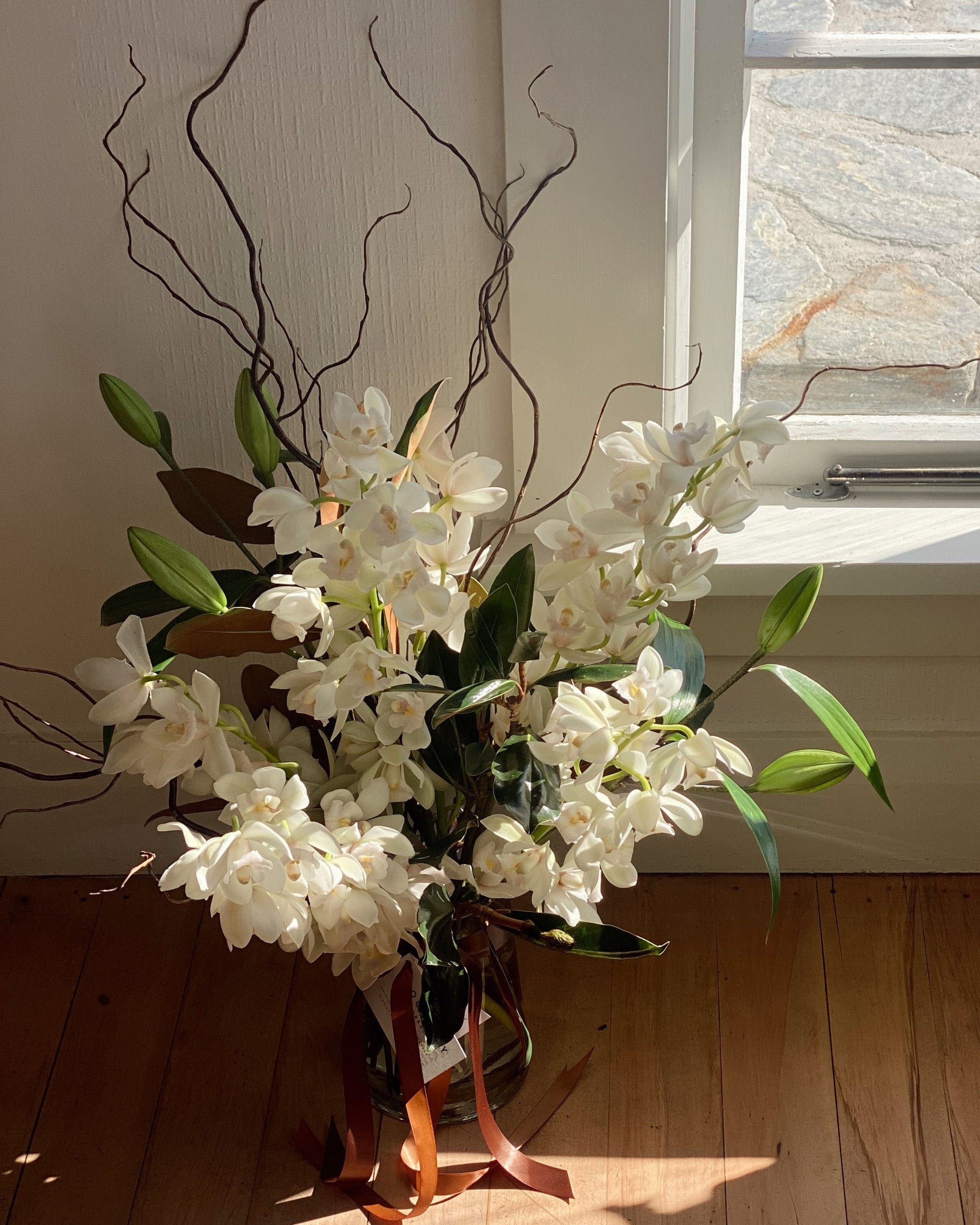 A glass vase from The Workroom NZ, containing the FRESH FLOWERS IN A VASE arrangement, showcases elegant white orchids and green leaves accented by curly brown branches. This floral display is illuminated by soft, natural light streaming through a nearby window, creating gentle shadows on the wooden floor.