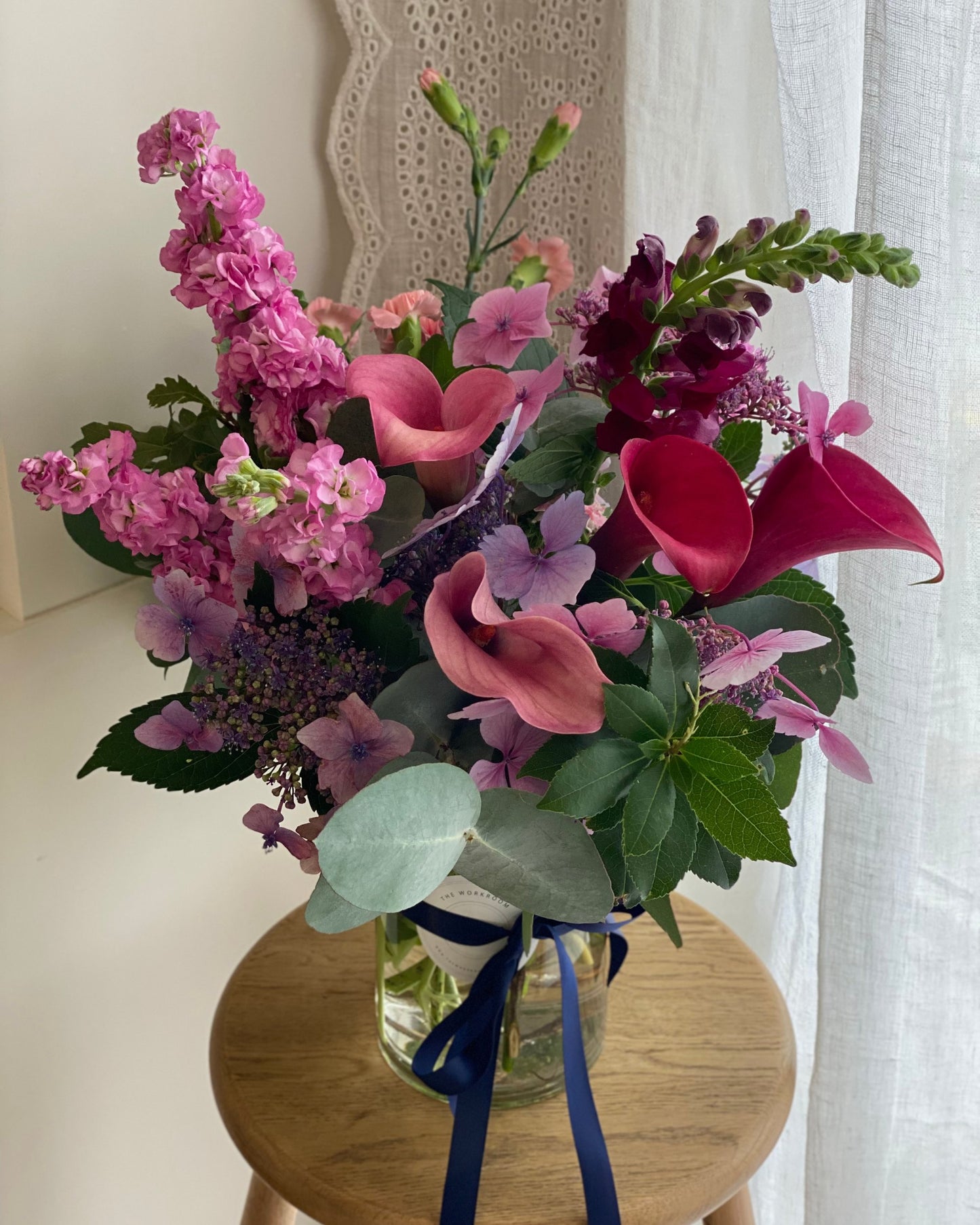 A vibrant bouquet from The Workroom NZ, featuring pink and purple seasonal blooms such as calla lilies and hydrangeas, is elegantly arranged in a clear glass vase with a dark blue ribbon. This beautiful display of "FRESH FLOWERS IN A VASE" is showcased on a wooden stool against lace curtains.