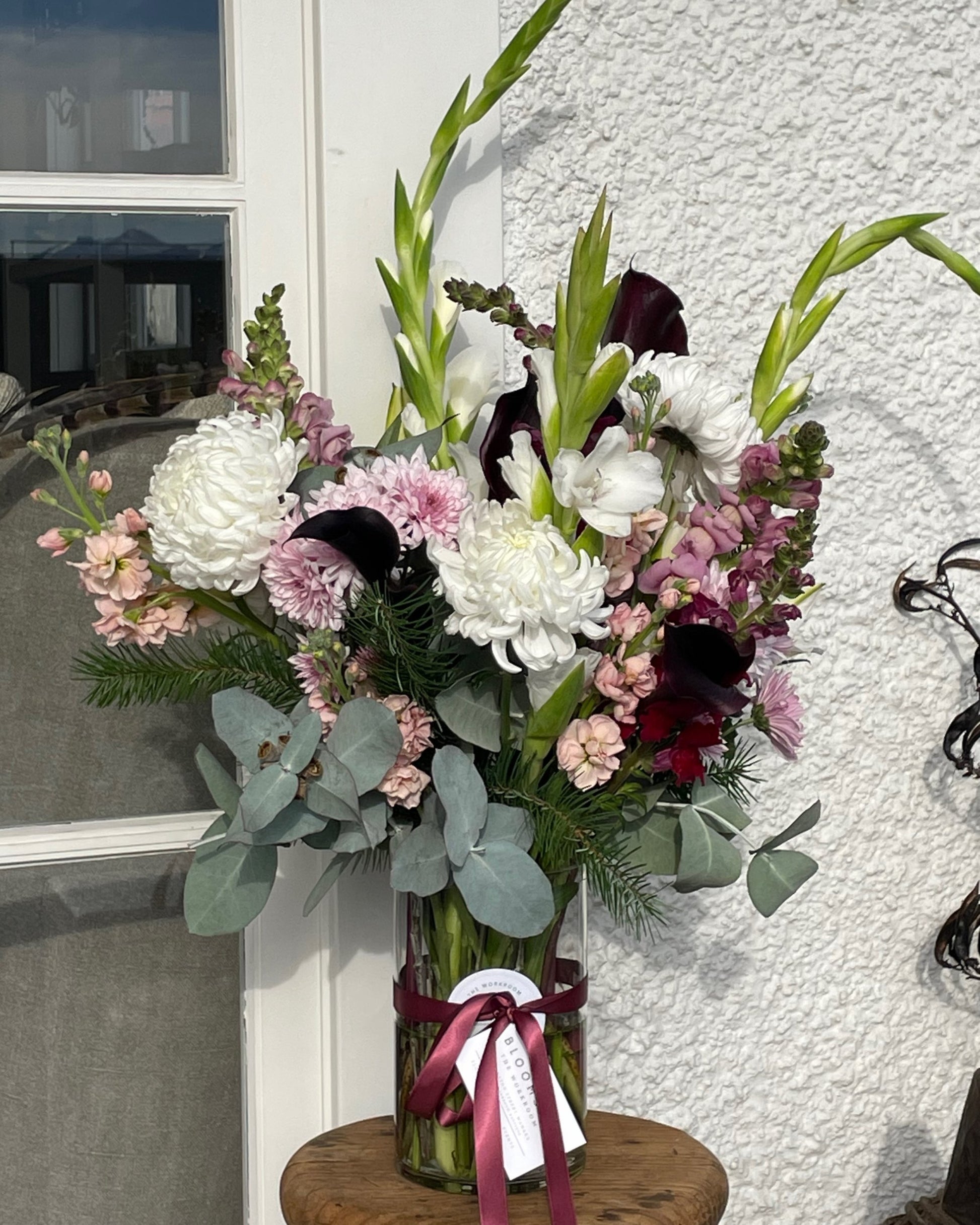 The Workroom NZ's "FRESH FLOWERS IN A VASE" features a tall and elegant bouquet in a glass vase with a burgundy ribbon, beautifully displaying white chrysanthemums, purple calla lilies, pink snapdragons, and green eucalyptus leaves. These seasonal blooms are artistically arranged against a textured white wall.