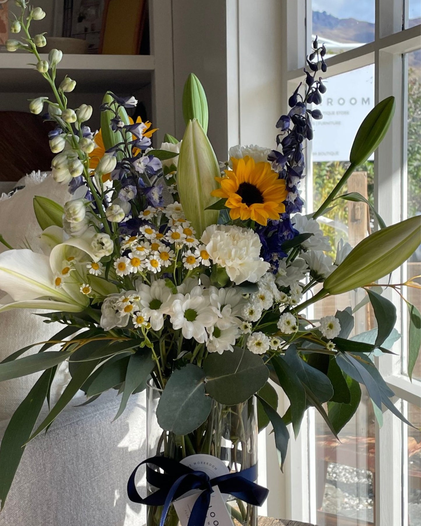 A bouquet of FRESH FLOWERS IN A VASE from The Workroom NZ features sunflowers, lilies, and vibrant seasonal blooms in white and purple tones. Sunlight shines on the arrangement near a window, with an elegant navy blue ribbon wrapped around the vase.