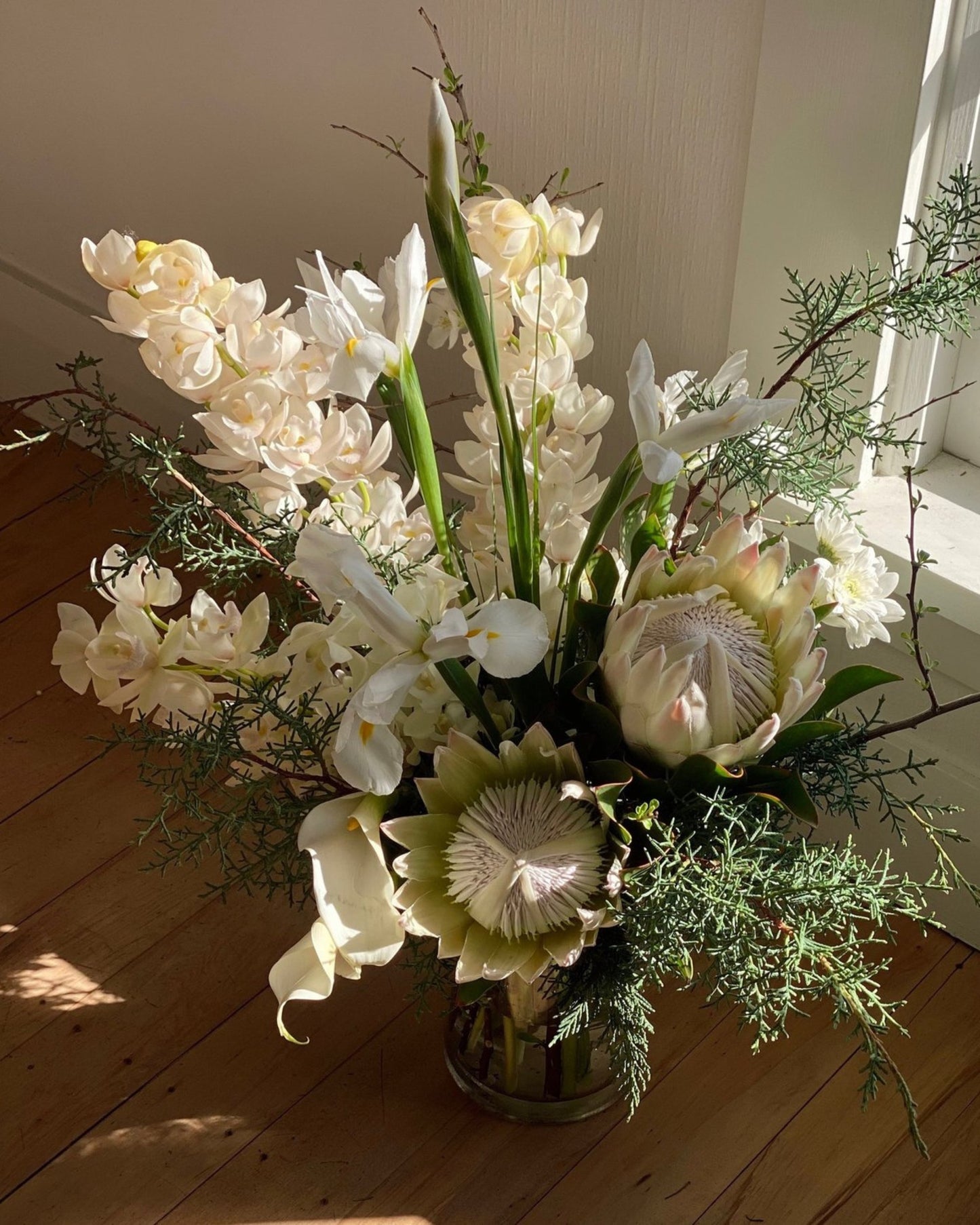 The Workroom NZ's FRESH FLOWERS IN A VASE showcases large protea flowers, white orchids, and sprigs of greenery in a glass vase. The arrangement is beautifully illuminated by natural sunlight on a wooden floor near a white wall, embracing seasonal blooms to add an elegant touch to any space.