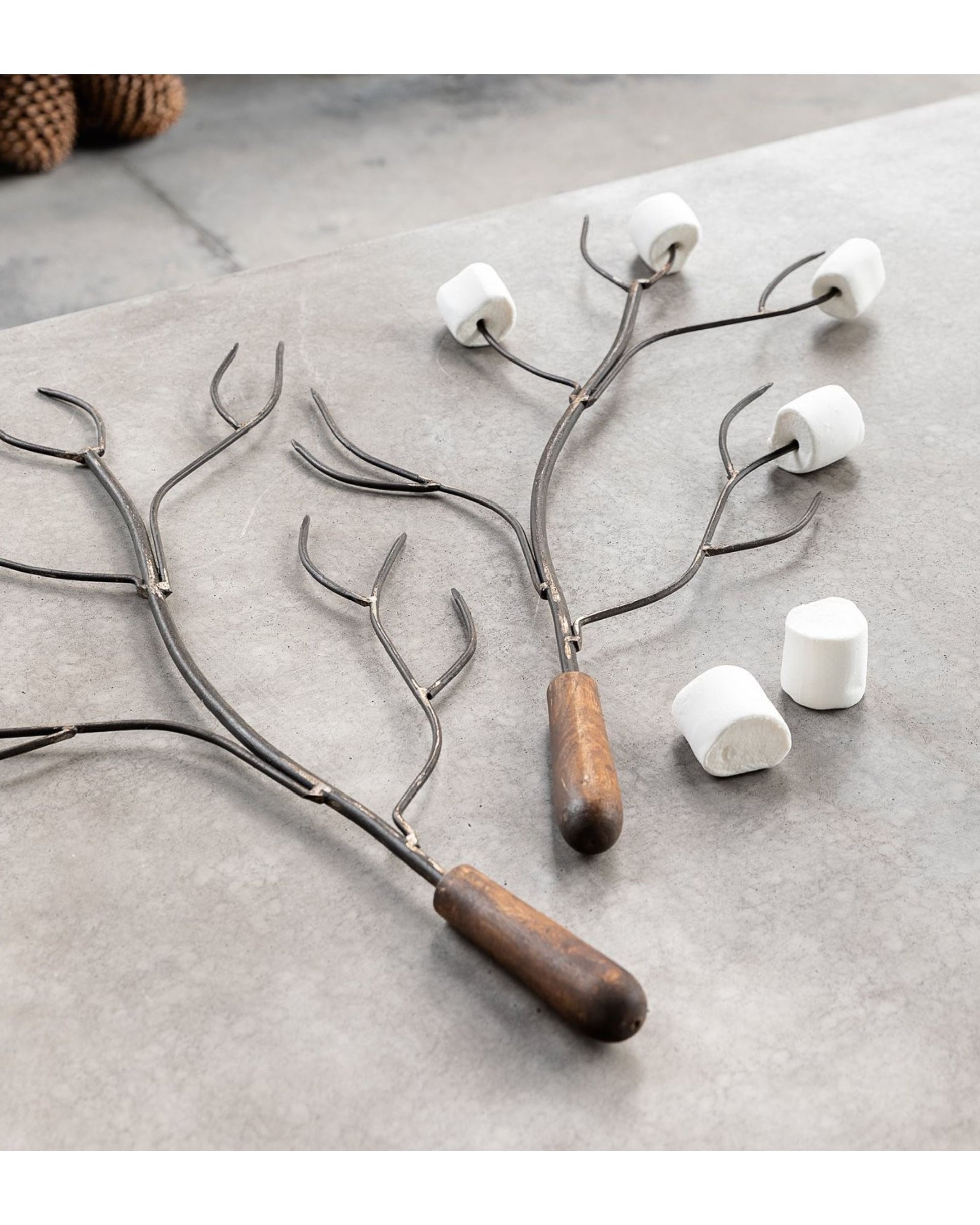 Two Hawthorne Sahar Marshmallow Forks with wooden handles rest on a table, their branch-like design lending a rustic touch. Handcrafted in India from reclaimed materials, each prong securely holds a marshmallow, while three extra marshmallows lie nearby, ready for roasting.