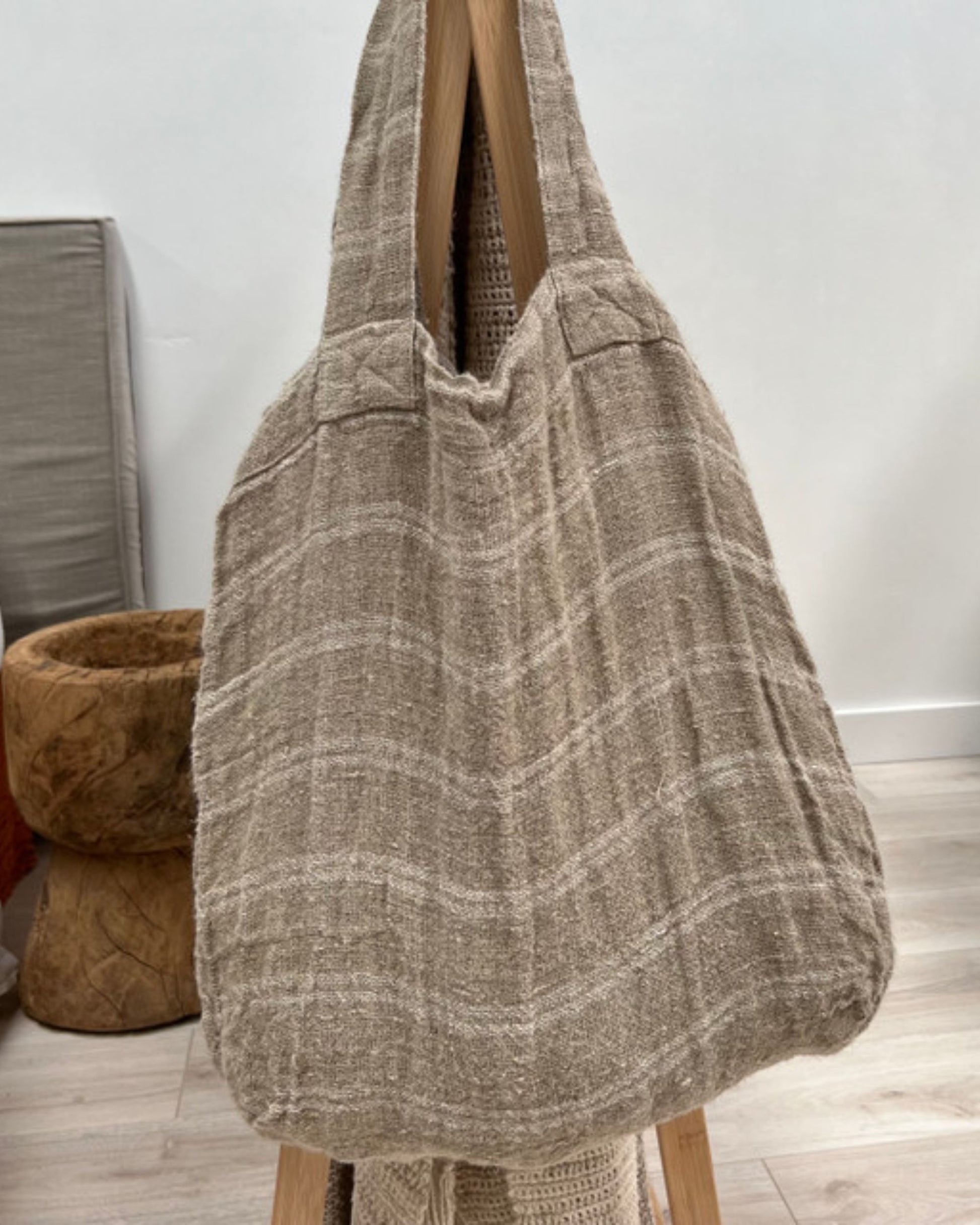 An ANGASTON HANDLOOMED LINEN TOTE BAG in white stripe by Rustic Linen hangs on a wooden stand. The background showcases neutral-toned furniture and a light wooden floor, enhancing the serene and natural ambiance.