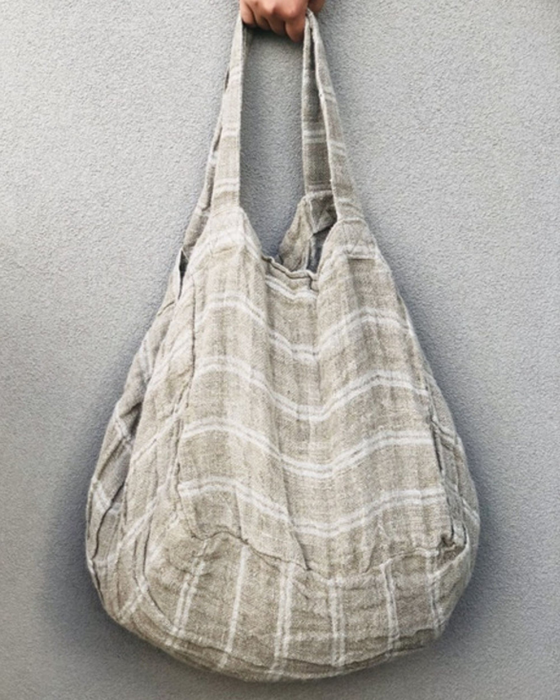 A hand is holding the ANGASTON ROUNDED LARGE TOTE BAG - WHITE STRIPE from Rustic Linen against a plain background. Crafted from pure linen, this tote bag features a soft, slouchy design with wide handles. Its subtle striped pattern enhances its rustic charm.