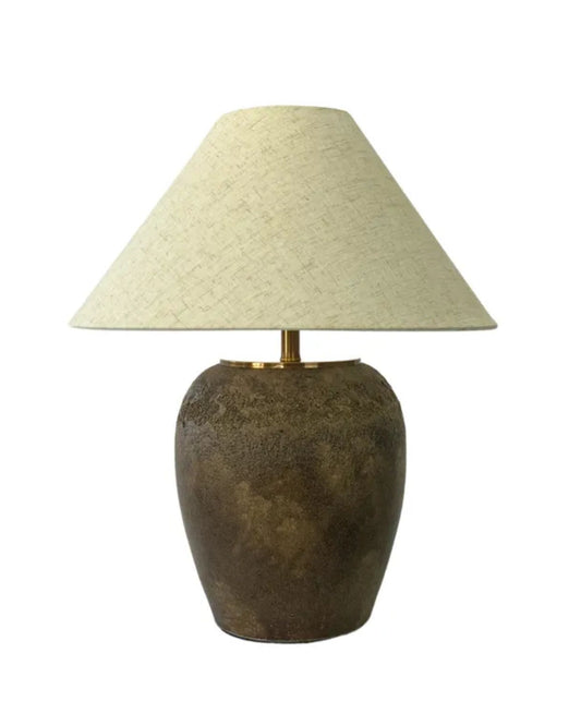 The CHELSEA CERAMIC TABLE LAMP by Capulet boasts a textured, brown ceramic base with an aged finish and is complemented by a wide, natural fabric shade. Its rustic charm is further enhanced by the elegant brass fitting that connects the shade to the base, set against a plain white background.