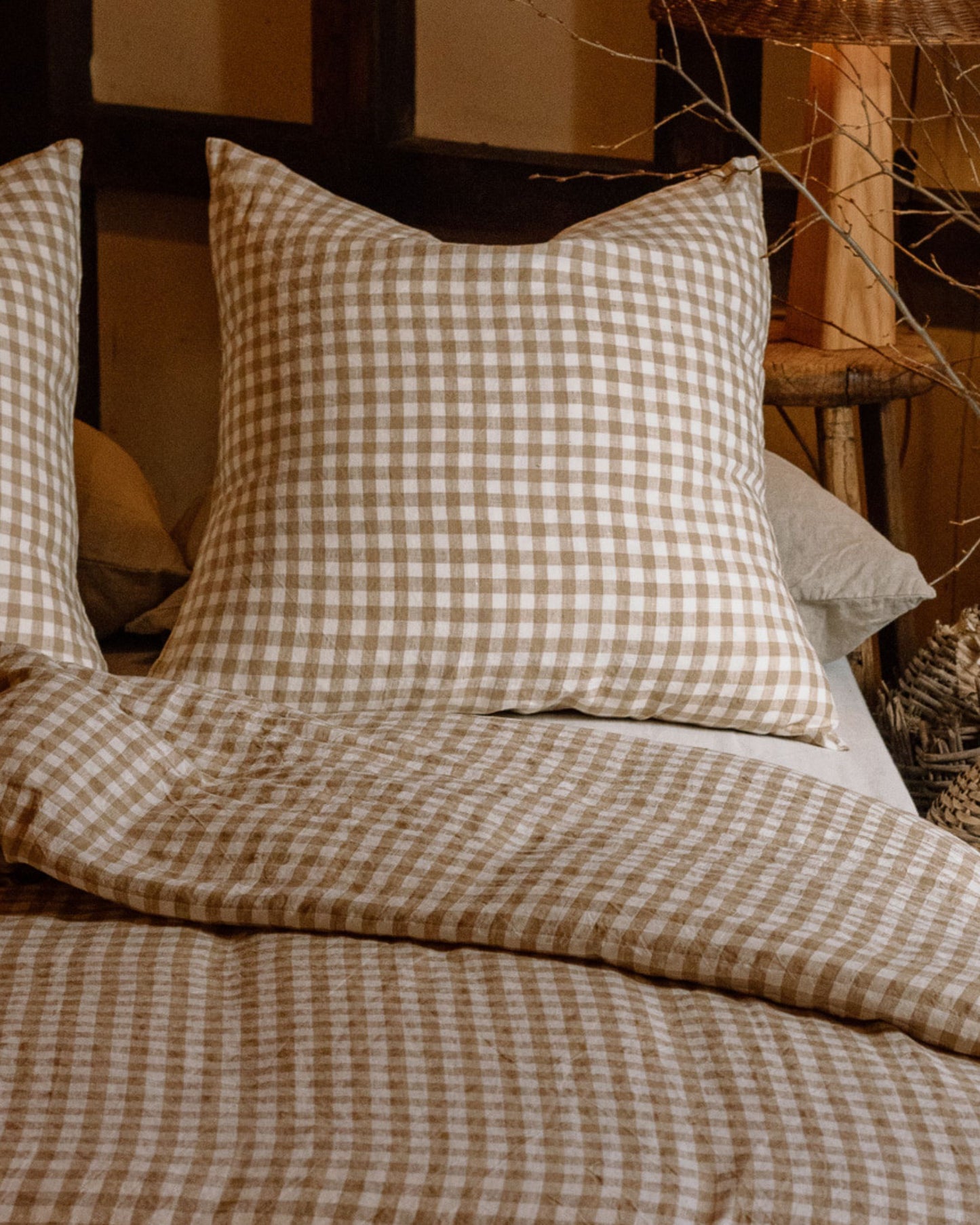 A comfortable bed is enhanced with Le Lin Euro Pillow Cases from The Workroom, featuring a cozy plaid pattern in earth tones. This pairs perfectly with a matching blanket made from 100% French flax linen bedding. The warm, rustic decor in the background creates an inviting and homey atmosphere.