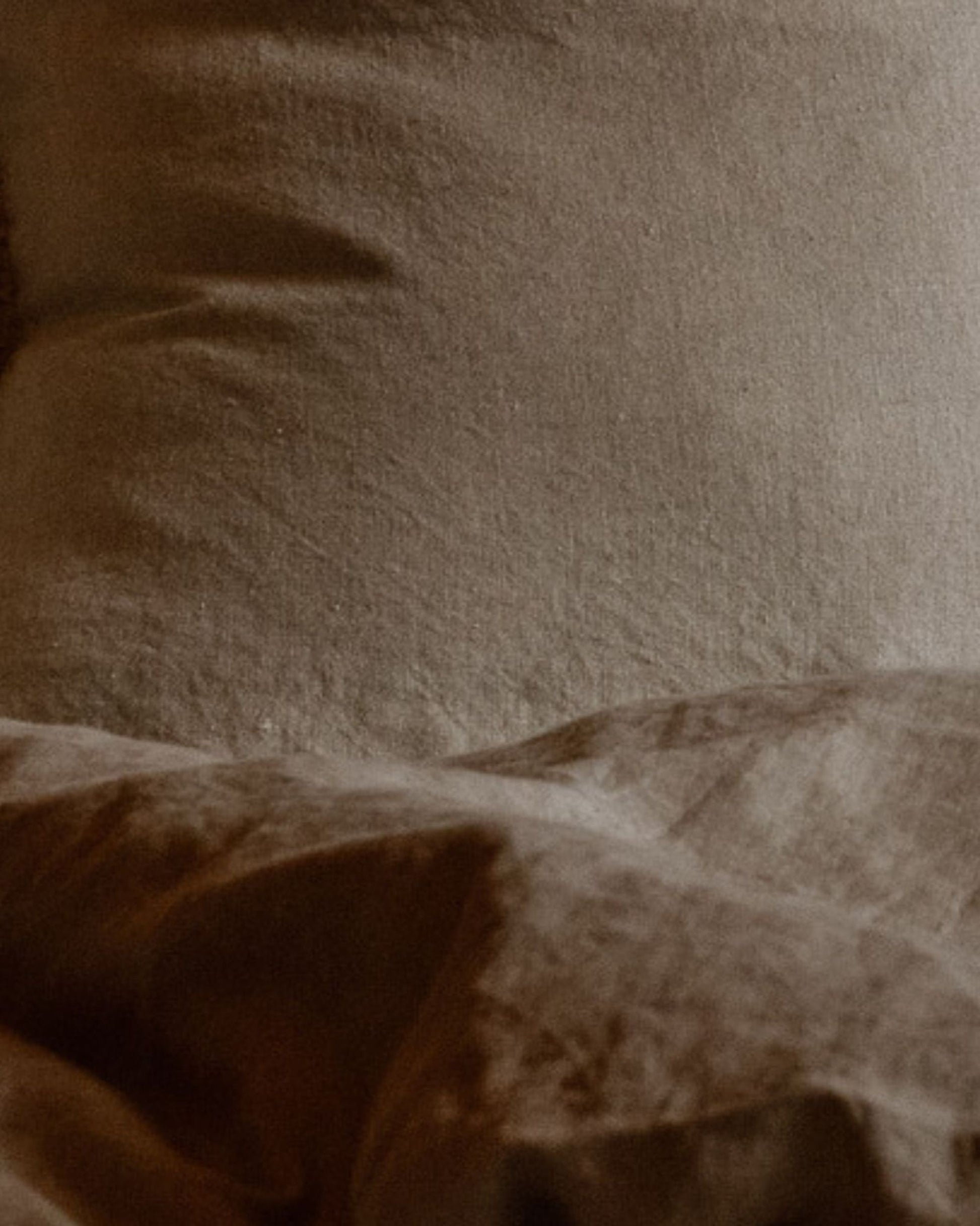 A close-up of a rumpled, beige LE LIN VALANCE pillow and blanket from The Workroom. The earthy-toned bedding boasts a soft, textured appearance, creating a cozy and relaxed atmosphere. Shadows add depth to the folds of the material.