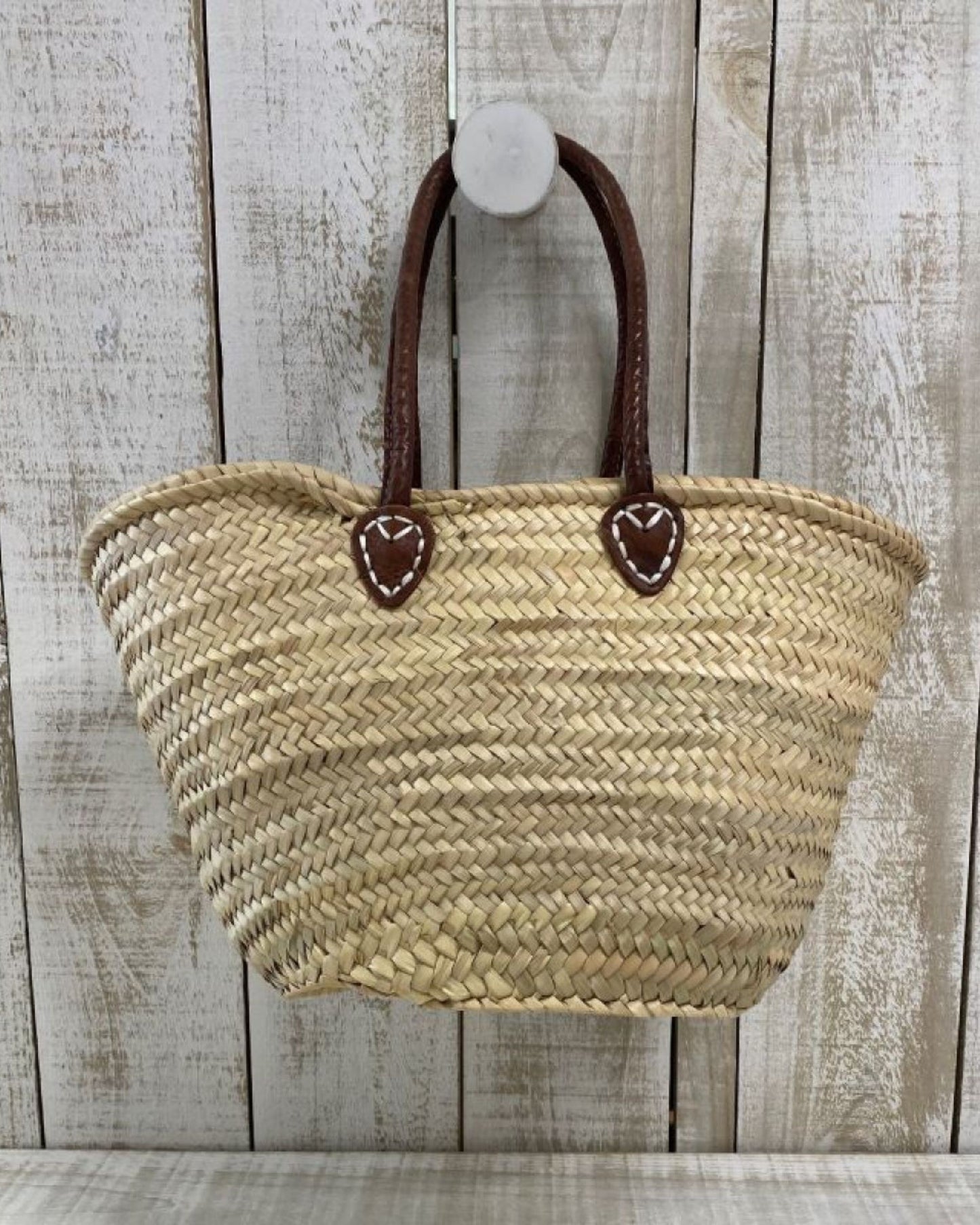 An Epicure Trader FRENCH MARKET BASKET WITH DELUXE HANDLE, featuring a woven straw design and dark brown leather handles, hangs on a wooden wall with a rustic, weathered appearance.