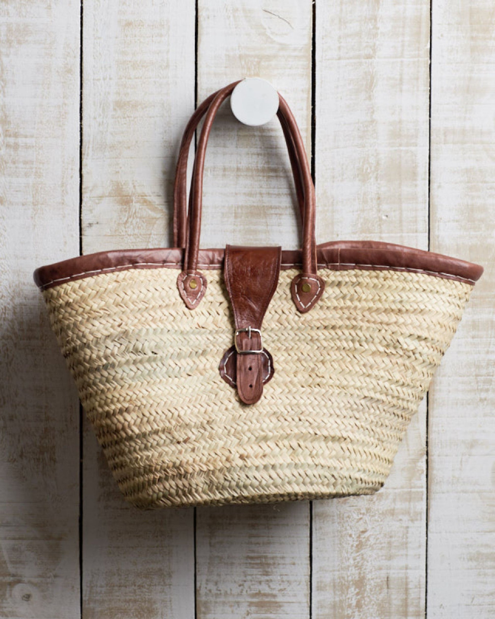 The FRENCH MARKET BASKET WITH BUCKLE from Epicure Trader, a distinctive Moroccan tote bag handwoven from palm fronds and featuring brown leather straps with buckle detail, hangs elegantly on a white wooden wall.