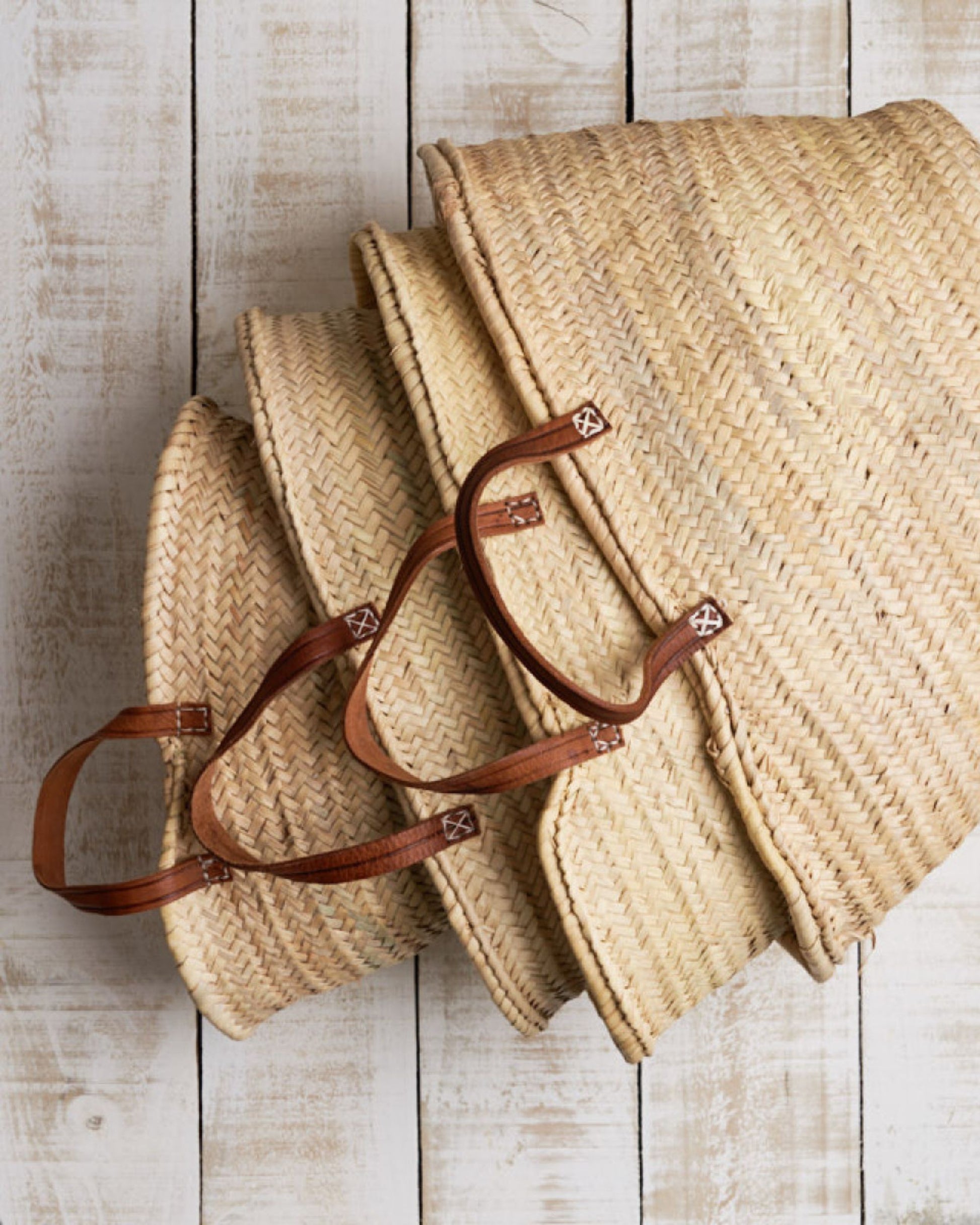 Three nested Epicure Trader FRENCH MARKET BASKET FLAT HANDLE made from woven straw, featuring hand-stitched brown leather handles, are displayed on a rustic whitewashed wooden surface. The baskets are arranged with the largest on the bottom and the smallest on top, highlighting their craftsmanship.