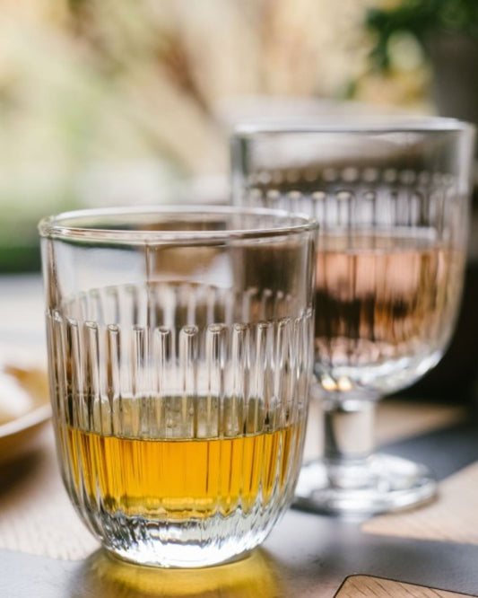 A close-up of two 290ml OUESSANT SHORT TUMBLERs by La Rochere on a wooden table. The glass in the foreground is filled with a golden liquid, while the one in the background contains a rose-tinted drink. Both dishwasher-safe glasses showcase a ribbed design, gently illuminated by sunlight.