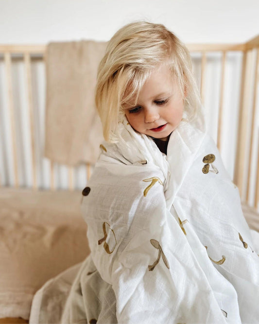 A young child with blonde hair sits wrapped in Leni's Muslin Swaddle Blanket - Mushroom, inside a crib. The background showcases a serene nursery setting with light-colored bedding and wooden crib railings, as the child appears both curious and peaceful.