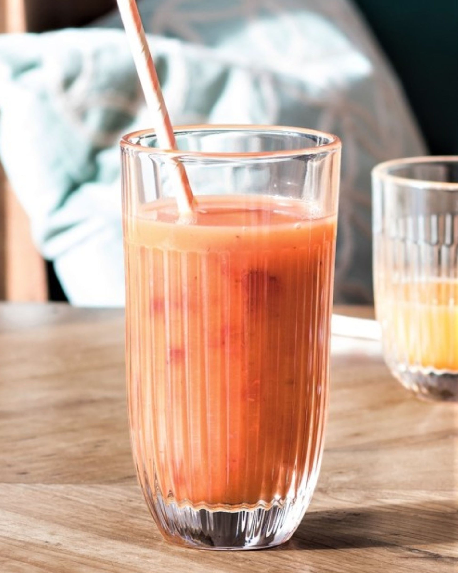 A tall OUESSANT TALL TUMBLER by La Rochere, filled with a pinkish-orange smoothie and accompanied by a straw, rests on a wooden table. In the background, another glass featuring a similar drink is partially visible. The scene is illuminated by natural light, highlighting the tumbler's dishwasher-safe design.