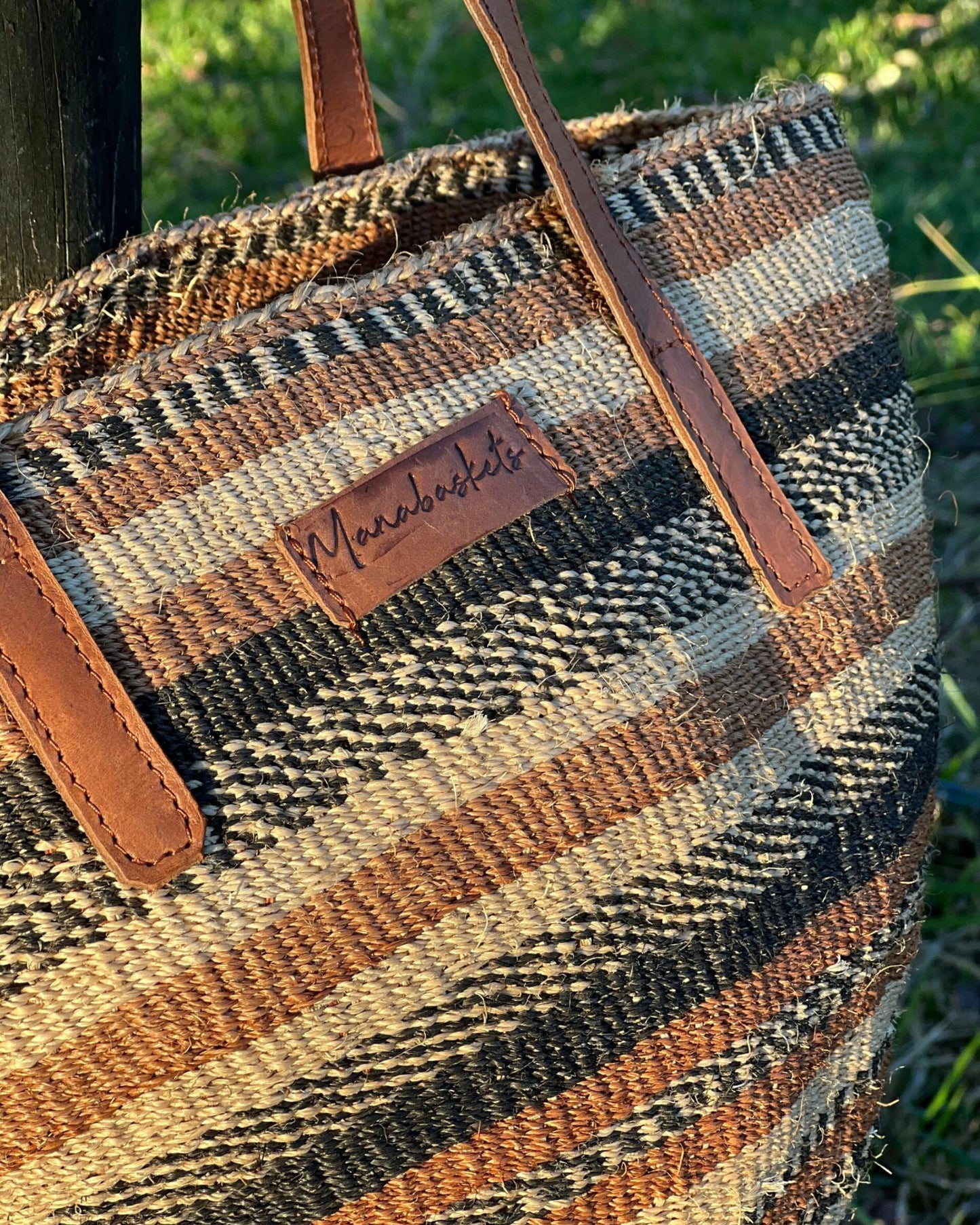 Close-up of the PENDA BASKET, a woven tote bag with brown, black, and beige stripes. The bag showcases leather shoulder handles and a stitched leather label displaying the brand name Manabaskets. Crafted by Kenyan women artisans, it rests on grass against a wooden post in the background.