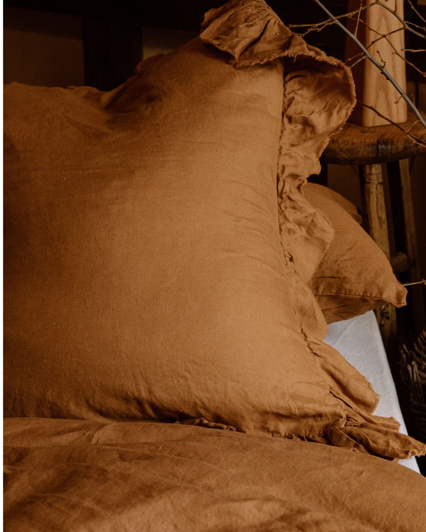 A cozy bed with soft, earth-toned pillows and a matching duvet in a rustic setting features The Workroom's LE LIN FRILLED EURO PILLOW CASES. Made from French flax linen, these pillowcases add elegance with their slightly crinkled texture and breathable fabric, creating a warm and inviting atmosphere.