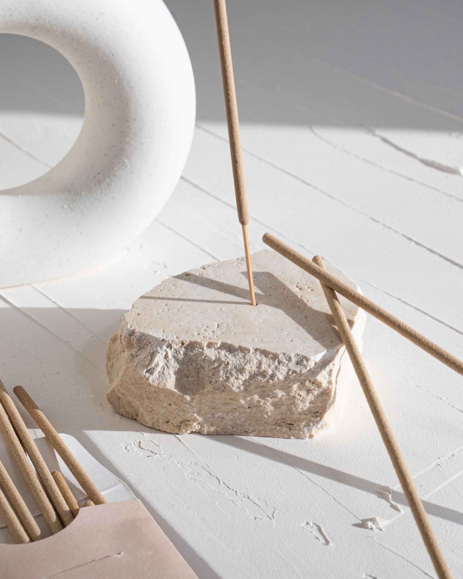 A handmade TRAVERTINE INCENSE HOLDER by Peggy Sue supports several burning incense sticks on a white wooden surface. The shadows from the incense enhance the calm and minimalist atmosphere, while a white curved object in the background complements the serene scene.