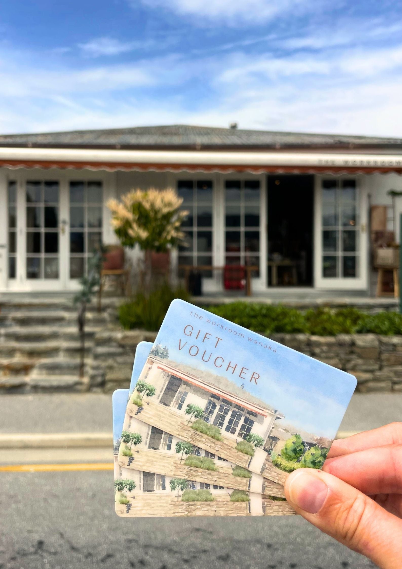 In a picturesque, rustic store featuring large glass windows, a hand holds three THE WORKROOM PHYSICAL GIFT VOUCHERS from The Workroom NZ. Each voucher reflects the charm of this delightful location, making it an ideal gifting option. The partly cloudy sky adds to the draw of an in-person shopping experience.