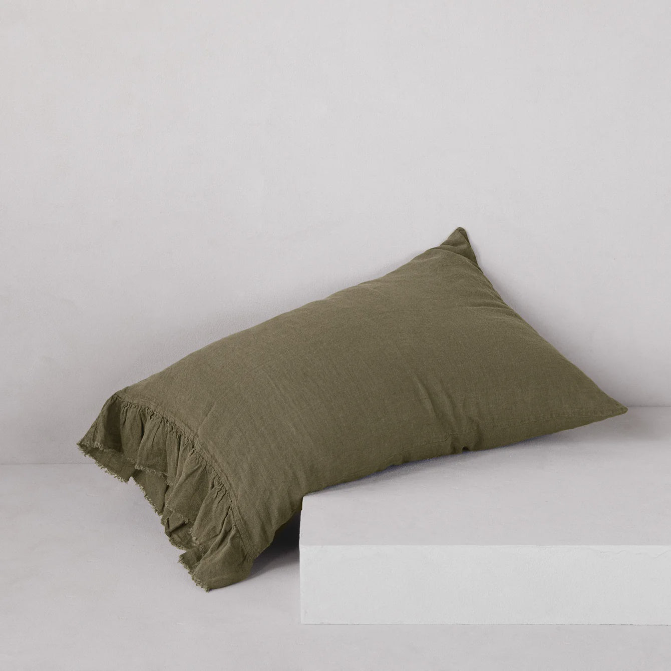 The HALE KRISTINE STANDARD PILLOWCASE by Hale Mercantile Co, in an olive green shade with frayed edges reflecting a touch of rustic romance, rests on a minimalist white block against a simple light gray background.