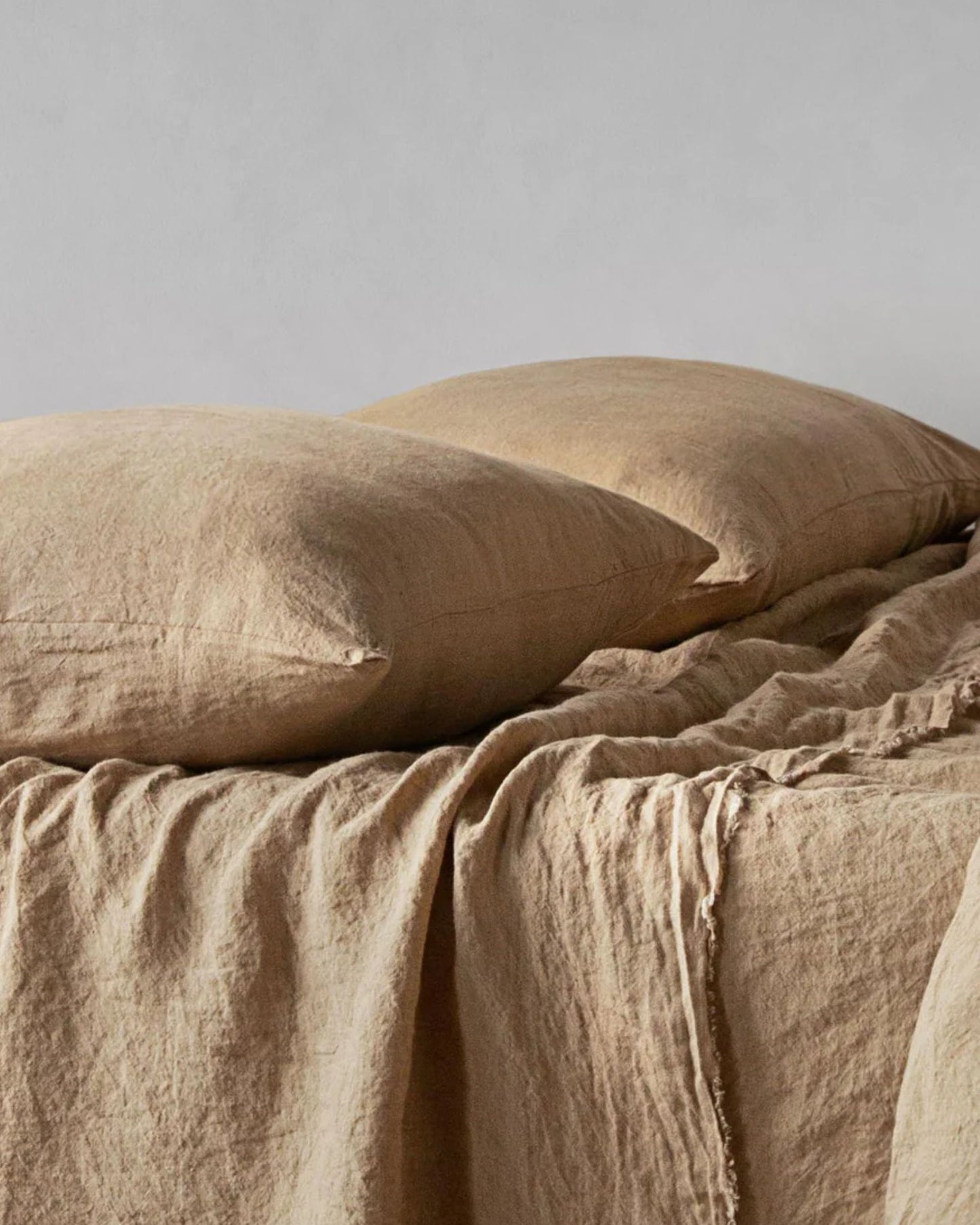 A neatly made bed with soft, beige linen bedding features the Hale Mercantile Co BASIX STANDARD PILLOW CASE and a slightly crumpled blanket against a neutral wall background.