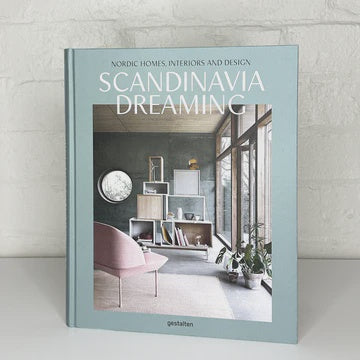 The image showcases the cover of the book "SCANDINAVIA DREAMING - GESTALTEN" by Flying Kiwi, emphasizing Scandinavian interiors and Nordic design. It presents a minimalist interior with expansive windows, a pink chair, and a shelf set against a white brick wall.