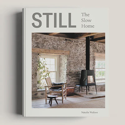 Cover of *STILL THE SLOW HOME* by Natalie Walton, from Nation Wide Books. This interiors book emphasizes sustainable living, showcasing a cozy room adorned with stone walls, a window, a wooden chair complete with a cushion, a small stool, and complemented by a modern fireplace.