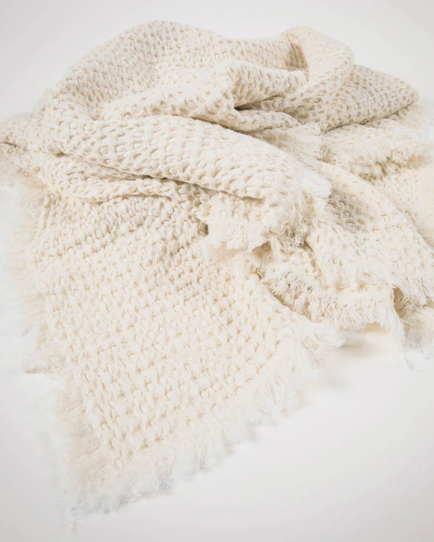 A detailed view of the ELEMENT THROW - IVORY by Eadie, showcasing its chunky weave and fringed edges. The throw blanket is elegantly arranged and softly illuminated, drawing attention to its plush, cozy texture with a subtle boho flair that enhances its appeal.