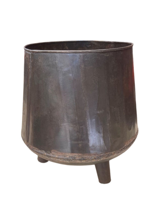 The ORIGINAL IRON THREE LEG PLANTER by Hawthorne is a weathered, dark metal planter featuring a wide opening and three short, sturdy legs. Its Indian origins are evident in its surface, which displays signs of age and rustic textures, contributing to its historic charm.