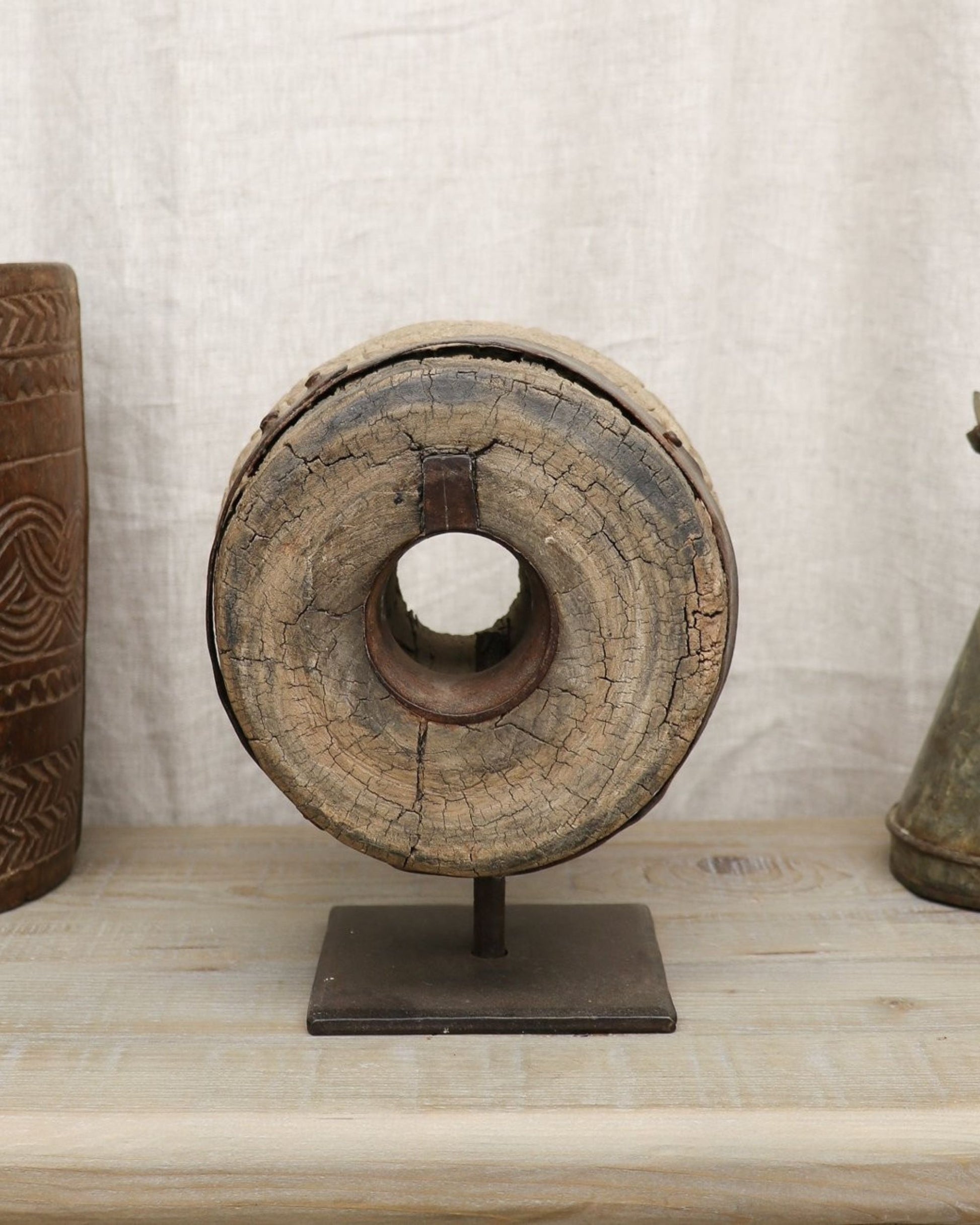 Displayed on a wooden surface among assorted shapes is the ORIGINAL WOOD SCULPTURE - SMALL by Hawthorne. This rustic, circular piece features a hollow center and is elegantly mounted on a metal stand. The weathered wood, adorned with visible cracks, exudes an antique charm reminiscent of India. Accompanying it are other intricately carved objects.