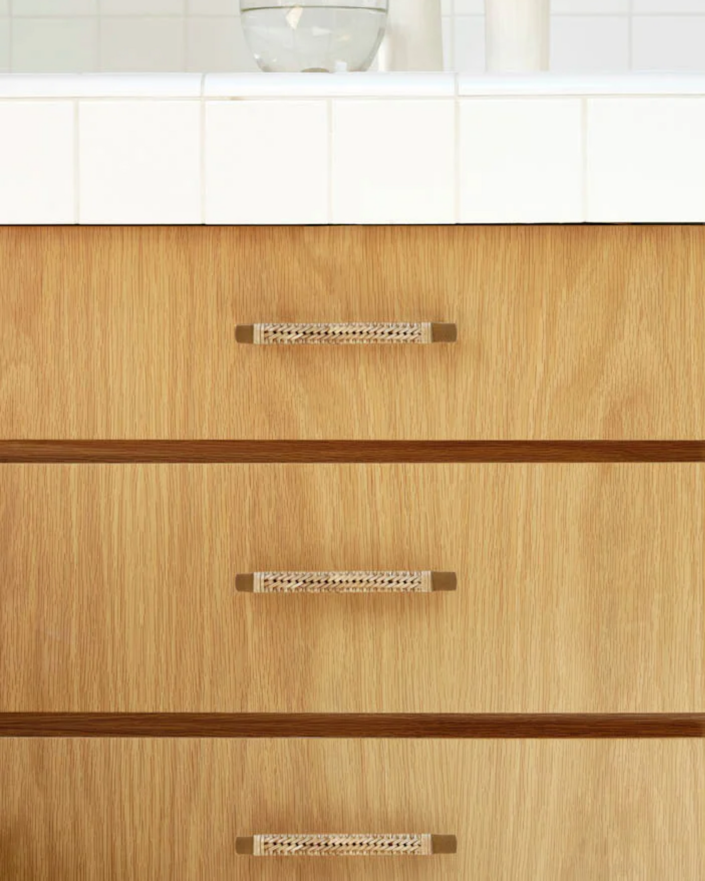 Close-up of a wooden dresser showcasing artisanal craftsmanship with three drawers, each featuring the 1954 DRAWER PULL by Society Ink. The top surface is a white tiled countertop, enhancing its aesthetic appeal, while part of a clear glass and a white object are visible.