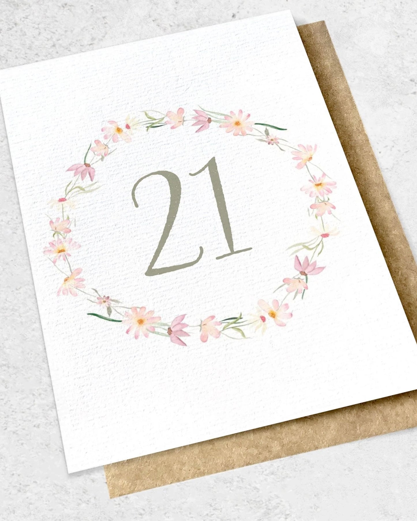 The Ink Bomb "21 - GREETING CARD" showcases a premium textured card featuring the number 21 at its center, surrounded by a delicate floral wreath of pink flowers on a soft background. This elegant card is paired with a classic kraft envelope.