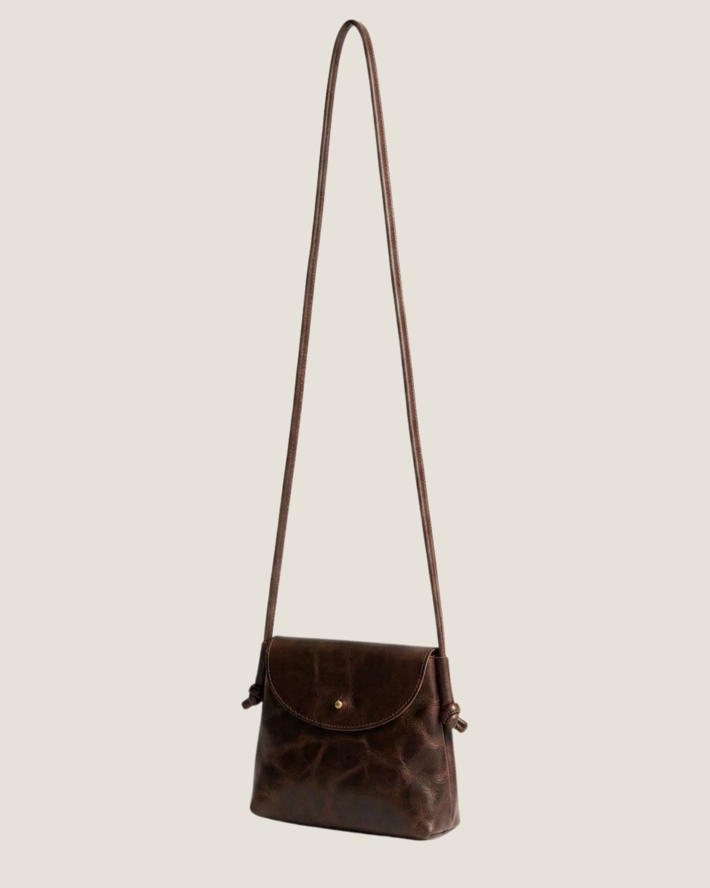 Image of the LEATHER KNOT PURSE by The Workroom, a minimalist black crossbody bag equipped with a long, slim strap. It showcases a small flap closure and features an attached tag on one side, made from full-grain leather for an elegant touch—making it a timeless statement accessory.