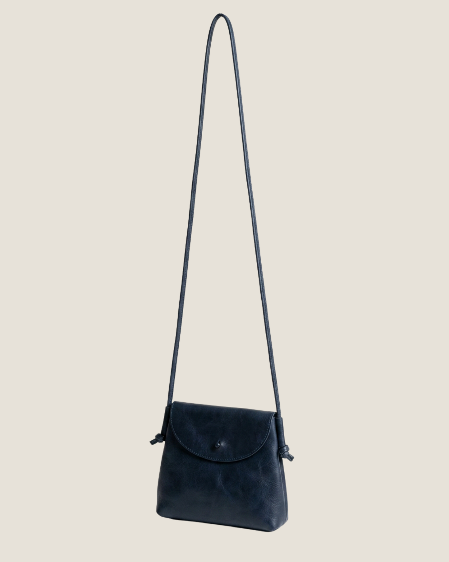 Image of the LEATHER KNOT PURSE by The Workroom, a minimalist black crossbody bag equipped with a long, slim strap. It showcases a small flap closure and features an attached tag on one side, made from full-grain leather for an elegant touch—making it a timeless statement accessory.