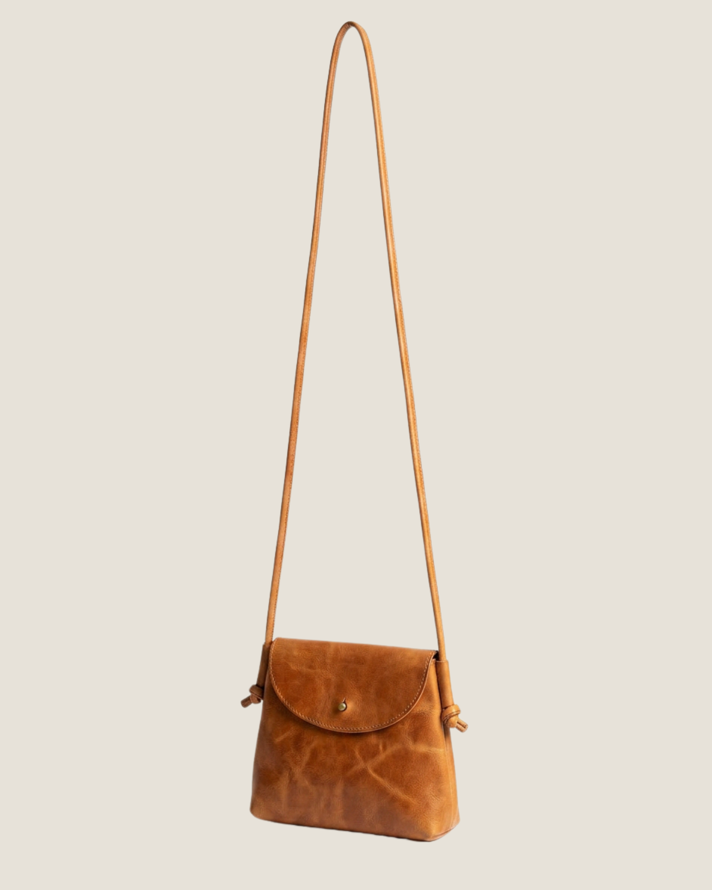 Image of the LEATHER KNOT PURSE by The Workroom, a minimalist black crossbody bag equipped with a long, slim strap. It showcases a small flap closure and features an attached tag on one side, made from full-grain leather for an elegant touch—making it a timeless statement accessory.