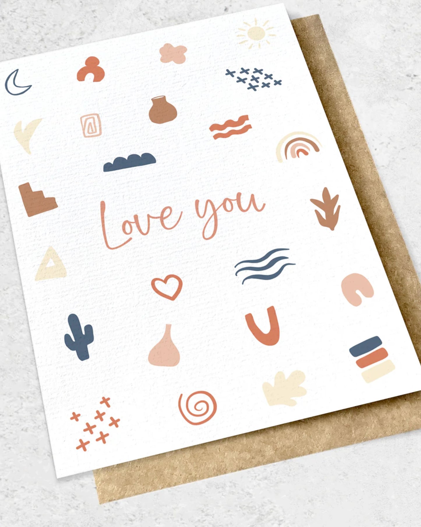 A premium greeting card from Ink Bomb, titled "LOVE YOU," showcases the phrase in the center, surrounded by abstract elements such as clouds, rainbows, and geometric shapes. It elegantly lies on a textured surface with a partially visible kraft envelope beneath it.