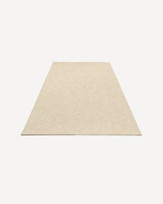 A beige, trapezoidal rug is laid flat against a neutral background, its shorter edge closer to the viewer, exuding minimalist charm. Nearby, a French Country RECTANGLE SHADE LINEN NATURAL 41X28CM adds a touch of nostalgia with its European fittings.