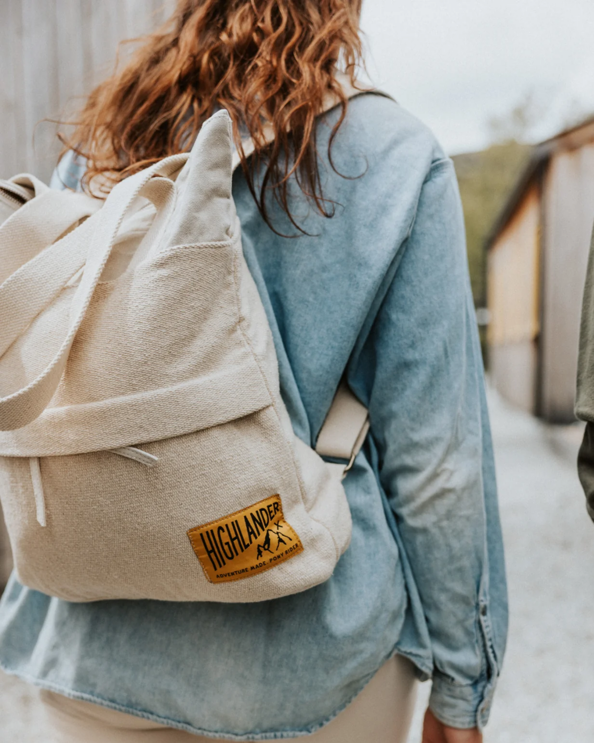 A woman with long hair wearing a denim shirt strolls outdoors, carrying the ADVENTURE BACKPACK - NATURAL by Pony Rider, crafted from upcycled truck canvas. The path is flanked by wooden structures, and a blurred figure moves beside her, highlighting the rugged charm of this handmade-in-India backpack.