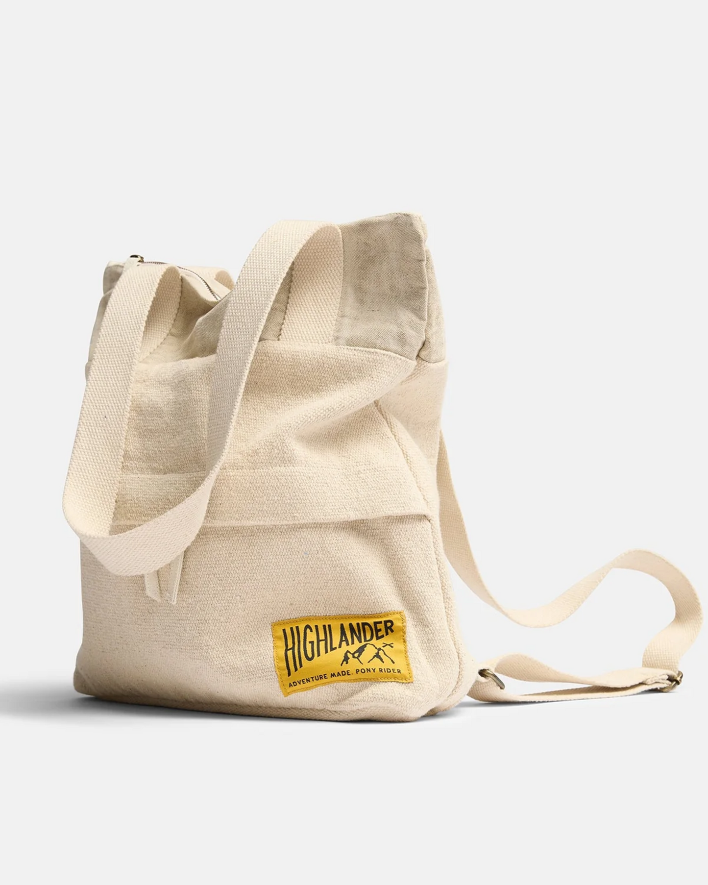 The ADVENTURE BACKPACK - NATURAL by Pony Rider is crafted in India from upcycled truck canvas. It features a long adjustable strap, a front pocket, and boasts an embroidered yellow patch with the text "Highlander Adventure Board Body Soul" accompanied by a mountain design.