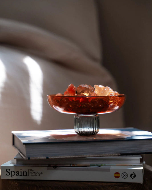 A glass bowl filled with decorative stones sits atop a stack of books, one of which is a travel guide about Spain. Sunlight softly illuminates the scene, casting gentle shadows, while an amber-hued bottle from the Amber Home Fragrance Set - Istanbul by Saarde adds a touch of elegance.