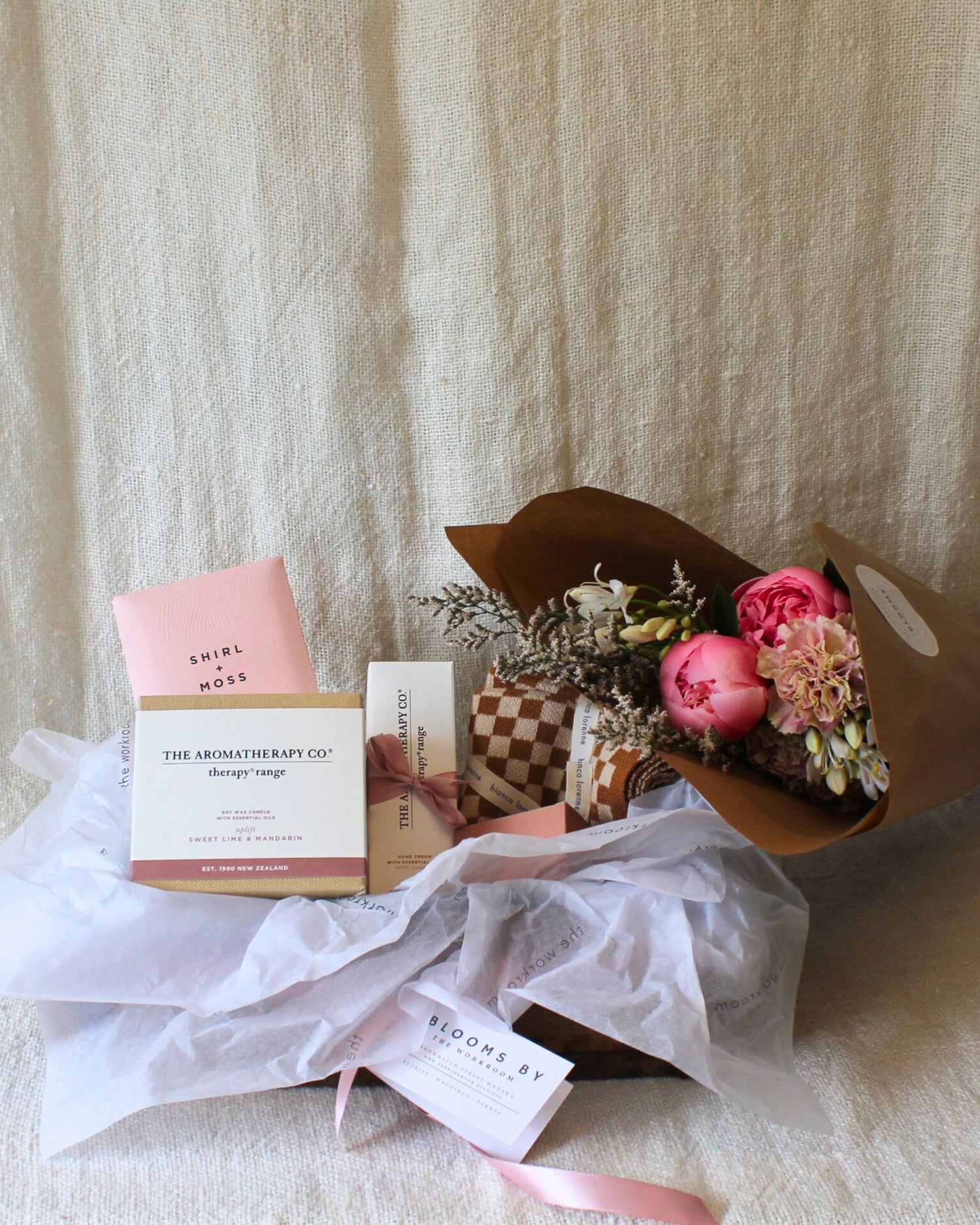 Introducing the AMORE BASKET - FOR HER by The Workroom NZ, a stunning gift basket that includes a tastefully arranged small bouquet of pink flowers wrapped in brown paper. This luxurious set also features an exquisite selection of Aromatherapy Co. and Bianca Lorenne skincare products, all elegantly packaged. Each item is nestled on white tissue paper against a soft beige background, radiating luxury and charm.