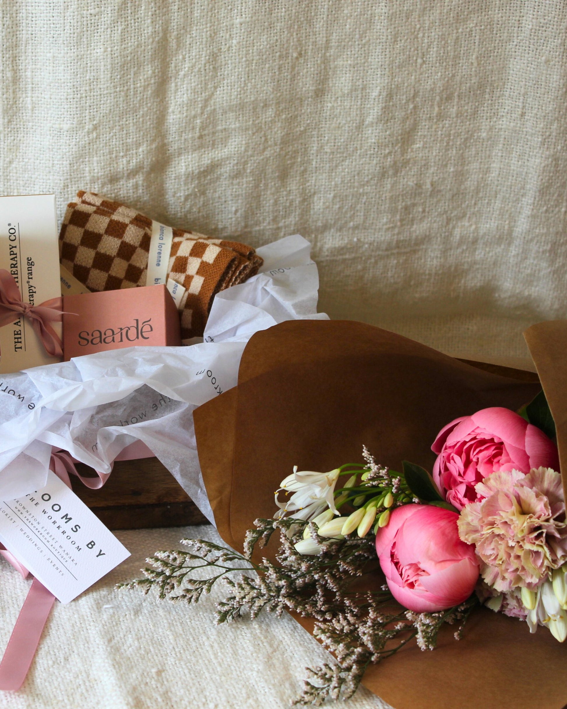 The AMORE BASKET - FOR HER, beautifully wrapped with a pink ribbon and featuring a checkered blanket branded with Saarde, sits alongside Bianca Lorenne's stunning bouquet of pink peonies and lush greenery, all elegantly arranged in brown paper set against a cream fabric backdrop. These exquisite offerings are brought to you by The Workroom NZ.