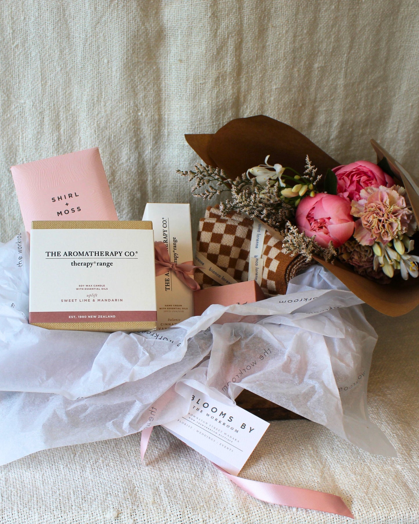 Explore the ideal gift option: the AMORE BASKET - FOR HER by The Workroom NZ. This exquisite arrangement includes a floral bouquet of pink peonies and white blossoms, complemented by aromatic products from Aromatherapy Co. and a rich chocolate bar. Each component is artfully displayed on soft fabric with pink and brown hues, echoing the signature style of Bianca Lorenne designs.