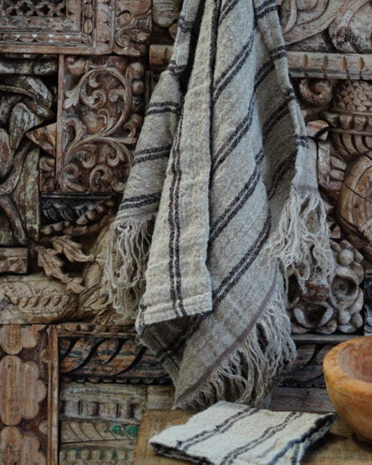 A Rustic Linen ANGASTON LINEN HAND TOWEL, featuring its textured beige fabric and dark stripes, hangs gracefully on an intricately carved wooden wall. A coordinating piece is folded neatly on the wooden surface in front, highlighting its superior linen quality and chic fringed edges.