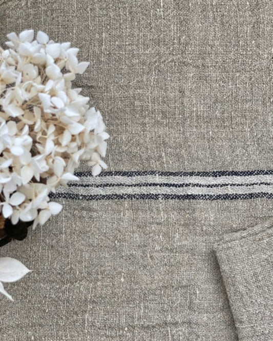 Close-up of the Rustic Linen ANGASTON BLUE STRIPE HANDLOOMED LINEN NAPKIN, showcasing its textured gray surface with black, white, and blue stripe detailing. A cluster of white flowers rests on the left side, providing a striking contrast to the fabric. A folded piece of the same material can be seen at the bottom right.