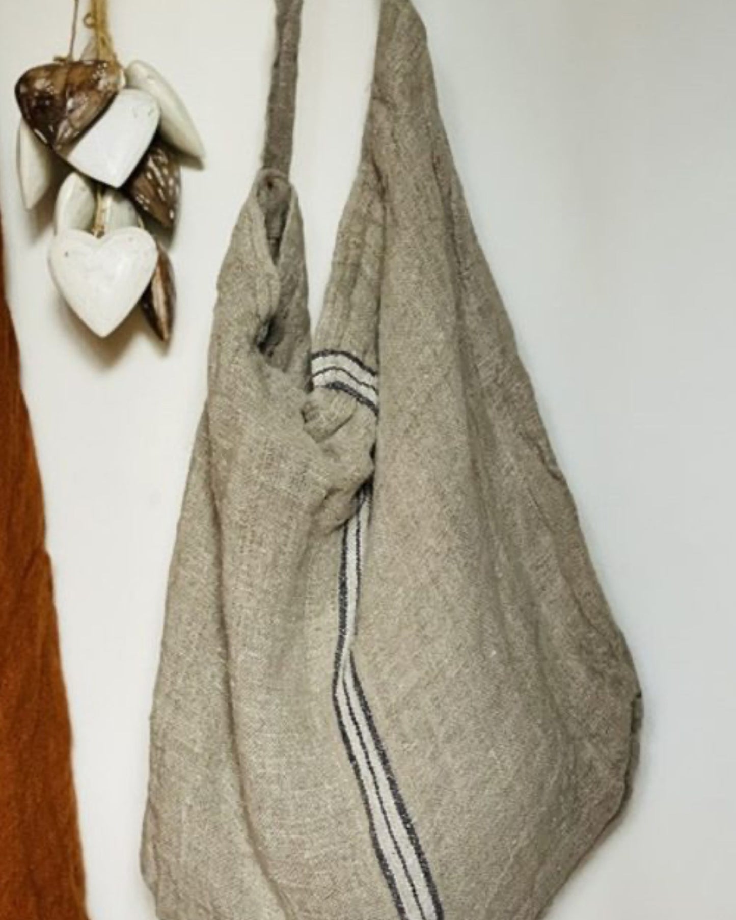 An ANGASTON HAND-LOOMED LINEN TOTE BAG from Rustic Linen, made from 100% pure linen with a navy center stripe, hangs elegantly on the wall. To its left, a string of white heart-shaped ornaments enhances the scene's charm. The neutral background beautifully accentuates the natural texture of this anti-allergenic accessory.