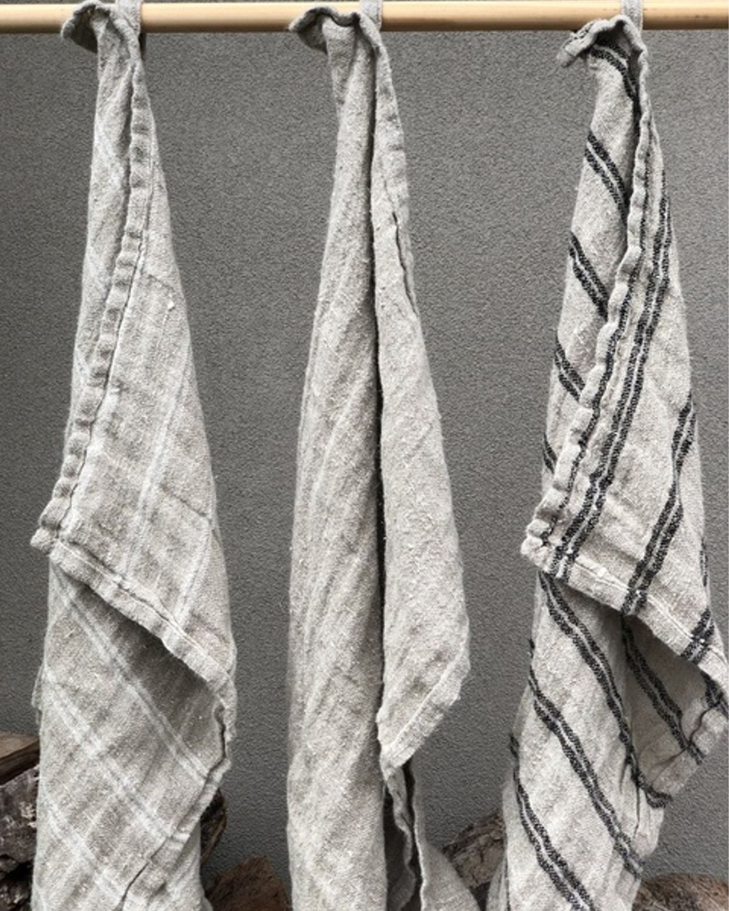 Three ANGASTON WASH CLOTHS WITH HANG LOOP from Rustic Linen hang from a wooden rod against a solid gray background. Crafted from premium linen, these wash cloths showcase varying patterns: the left one features a subtle checkered design, the middle displays faint diagonal stripes, and the right boasts bold black stripes.