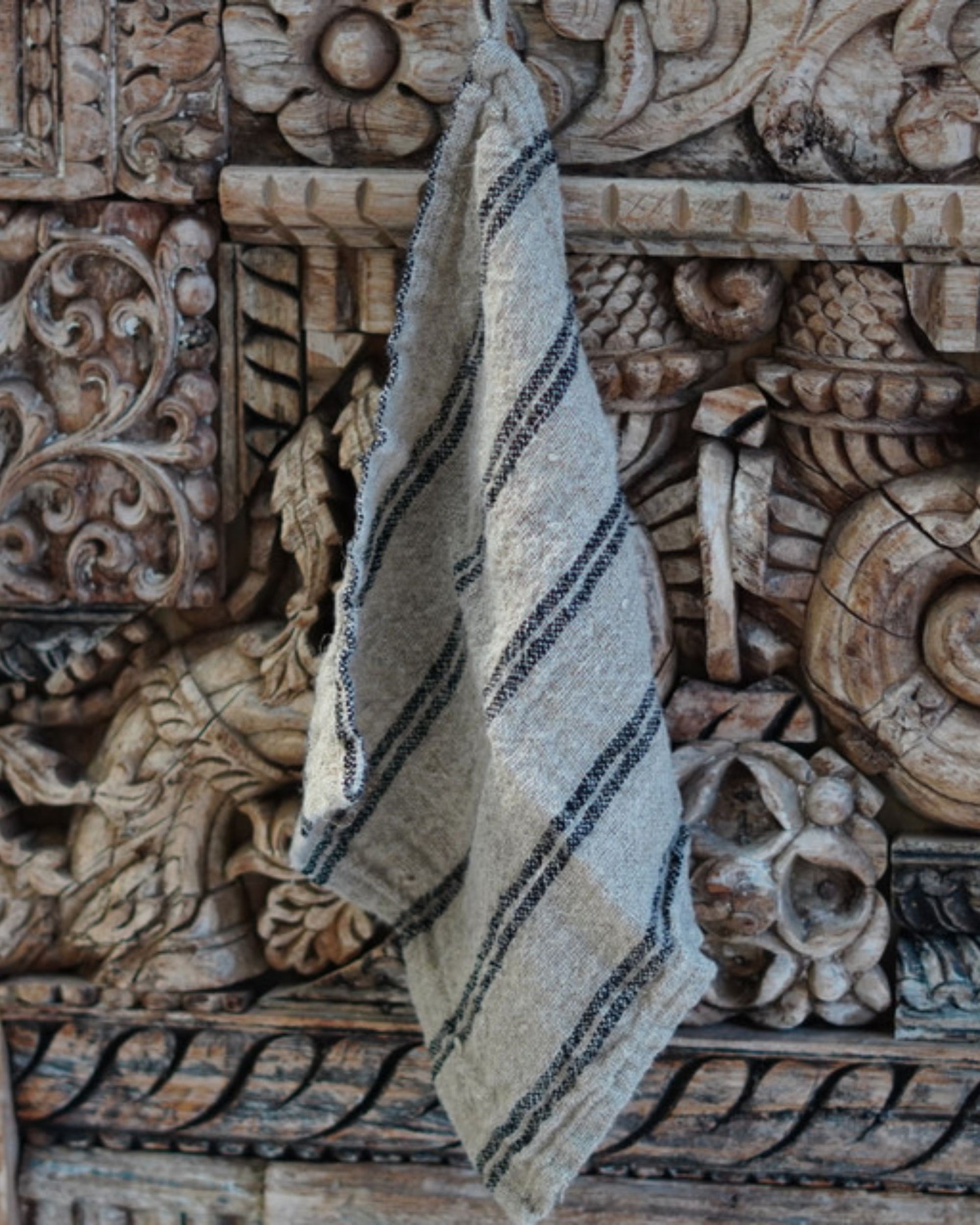 An ANGASTON WASH CLOTH WITH HANG LOOP from Rustic Linen, featuring a textured, striped design reminiscent of premium linen, drapes over ornate wooden panels intricately carved with floral and geometric patterns. The weathered wood showcases exquisite craftsmanship.