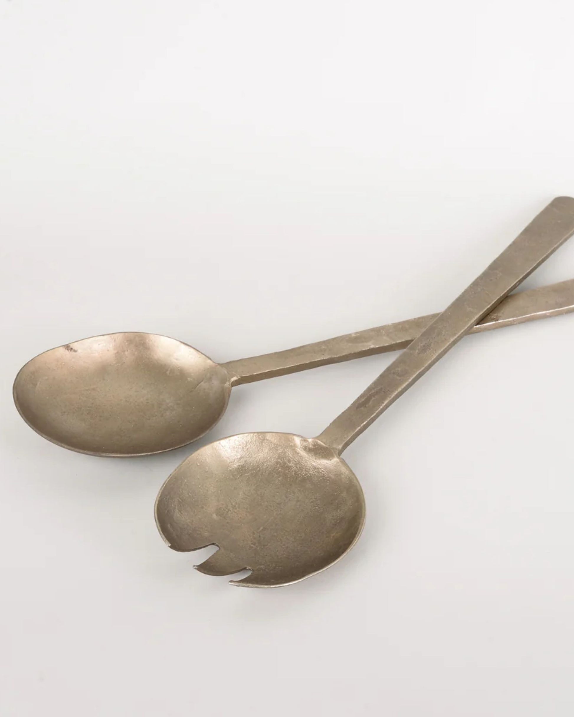 Two rustic, handmade ANTIQUE FLAT HANDLE SERVING SPOONS by New Direction are set against a plain background. One showcases a smooth, rounded shape, while the other resembles a spork with its serrated edge. These vintage utensils feature simple, elongated handles that exude an antique charm.