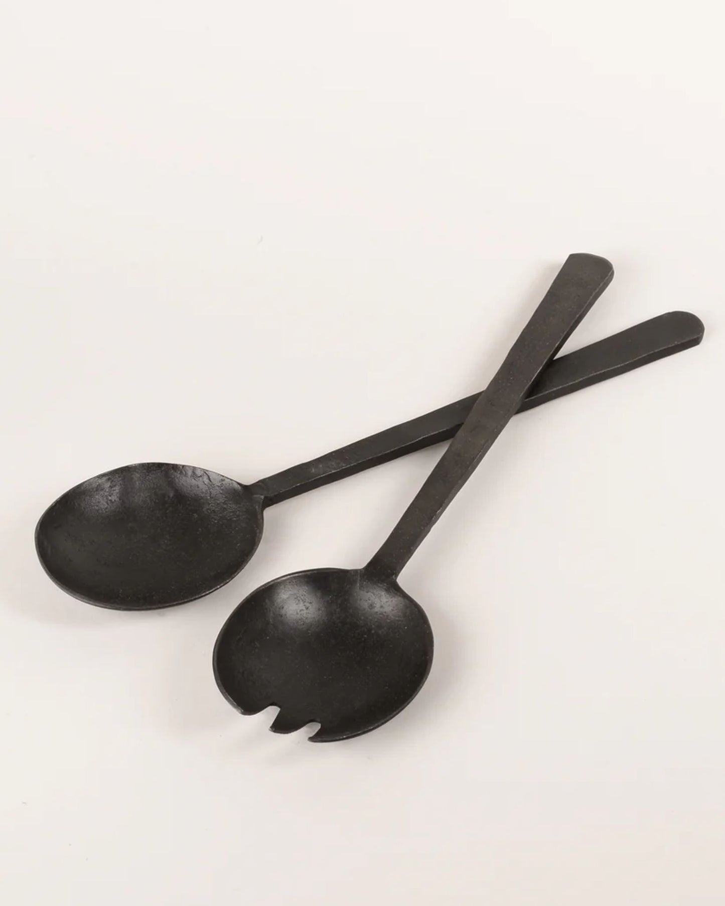 Two ANTIQUE FLAT HANDLE SERVING SPOONS from New Direction, with a black metal finish reminiscent of vintage serving utensils, are crisscrossed on a light surface. One spoon features a classic design while the other is adorned with fork-like prongs, combining style with functionality.