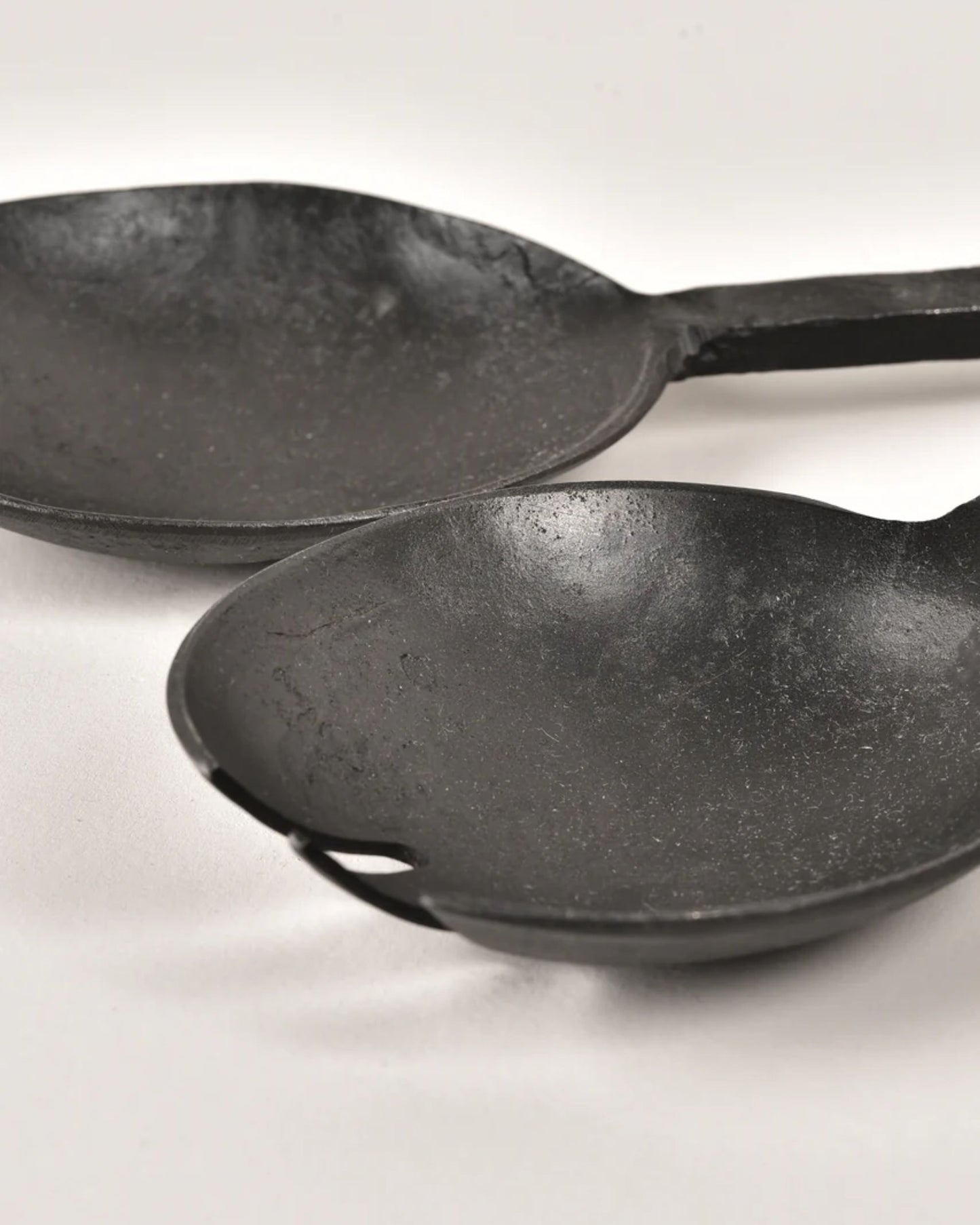 Close-up of two ANTIQUE FLAT HANDLE SERVING SPOONS by New Direction, featuring black cast iron with textured surfaces, placed side by side on a white background. These vintage serving utensils have a rustic appearance and seem well-used, evoking memories of eras gone by.